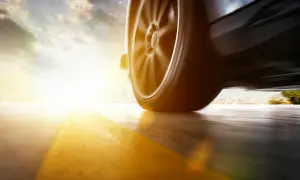 Roues d'une voiture en mouvement, le soleil rasant inonde la scène d'une lumière dorée, floutant le paysage urbain au loin.