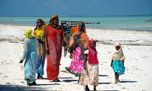 Kobiety i dzieci w kolorowych strojach spacerują po plaży Zanzibaru. Przed wyjazdem warto sprawdzić zalecenia dotyczące zanzibar szczepienia.