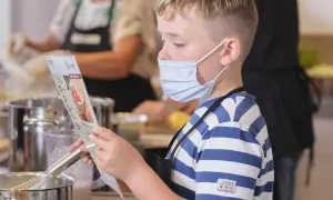 Junge mit Maske liest Rezept, während andere kinder kochen selbst. Küchenutensilien und Töpfe sind zu sehen.