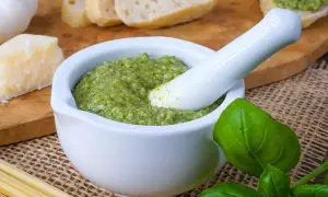 Frisches Pesto in einem Mörser, bereit zum Haltbarmachen mit Salz. Daneben liegen Brot, Parmesan, Knoblauch und Pasta.