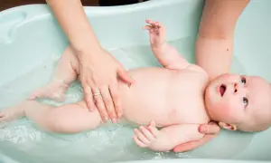 Rączki mamy delikatnie trzymają dziecko w wodzie w wanience dla niemowlaka. Maluch z zaciekawieniem patrzy w górę.