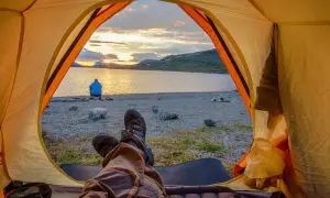 Blick aus dem Zelt auf einen See bei Sonnenuntergang. Eine Person sitzt am Ufer. Ja, darf man in Schweden wildcampen!