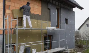 Pracownik montuje wełnę mineralną na ścianie domu, porównując jej grubość ze styropianem.