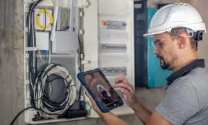 Technik w kasku i okularach ochronnych wykonuje przegląd instalacji elektrycznej, analizując dane na tablecie.