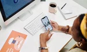 Une développeuse consulte son blog sur son téléphone, entourée d'un ordinateur, d'une tasse de café et de documents de conception.