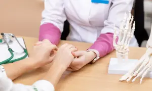 Arzt hält Patientin die Hand. Modelle von Fuß und Hand liegen auf dem Tisch. Hoffnung auf eine Wunderwaffe gegen Arthrose.