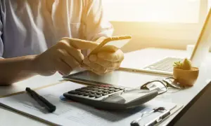 Osoba korzysta z telefonu, planując rezygnację z PPK. Na biurku kalkulator, laptop i dokumenty.