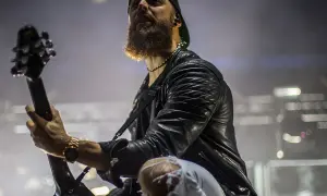 Charyzmatyczny gitarzysta z brodą podczas koncertu Bullet for My Valentine, w skórzanej kurtce i podartych spodniach, gra na gitarze.
