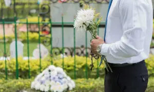 Mężczyzna w białej koszuli i niebieskim krawacie trzyma bukiet białych kwiatów i różaniec. To jego urlop okolicznościowy śmierć bliskiej osoby.