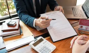 Biznesmen prezentuje umowę kredytową klientowi. Na stole leżą kalkulator, model domu i klucze.