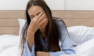 Een vrouw ligt moe in bed, haar hand voor haar ogen. Ze draagt een blauwe pyjama.