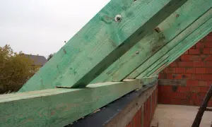 Zielone krokwie dachowe połączone na długości, tworzące konstrukcję nad ceglanym murem.