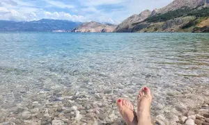 Stopy w klapkach na kamienistej plaży, krystalicznie czysta woda i górzyste wybrzeże. Raj na wyspie Krk.