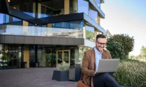 Uśmiechnięty mężczyzna w okularach pracuje na laptopie przed nowoczesnym budynkiem. Może właśnie sprawdza swój dodatek mieszkaniowy.