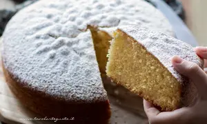 Una fetta di torta margherita soffice e profumata, cosparsa di zucchero a velo, viene sollevata da una mano.