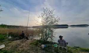 Dwóch wędkarzy siedzi nad jeziorem Wdzydze, czekając na branie ryb. Słońce zachodzi, malując niebo na złoto.