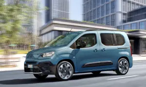 Un Fiat Doblò 9 posti blu metallizzato sfreccia in città, con grattacieli moderni sullo sfondo.