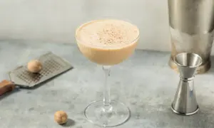 Ein cremiger Brandy Alexander Cocktail in einem Coupette-Glas, garniert mit Muskatnuss, neben einem Schnapsmaß und Muskatnüssen.