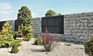 Nowoczesne ogrodzenie gabionowe montaż: kamienne kosze z czarnymi panelami, otoczone zielenią i kamyczkami.