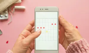 Aplikacja na telefonie pokazuje kalendarz cyklu. Czy można iść na wizytę do ginekologa podczas okresu? Na zdjęciu widać też tampony i tabletki.