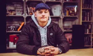 Młody mężczyzna w czapce i kurtce, z tatuażami na rękach, siedzi przy drewnianym stole. Wygląda jakby przeżywał swój **frosti wiek**, pełen pasji i determinacji.