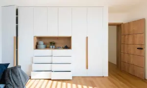 Armarios ocultos en pared blancos con nicho de madera y cajones abiertos. Puerta de madera a la derecha.