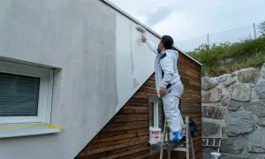 Mann streicht Fassade ohne Reinigung mit Roller. Ein Eimer Farbe steht bereit.