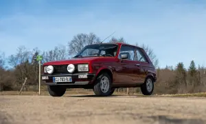 Röd Peugeot 104 med extraljus parkerad på en grusväg, med träd och blå himmel i bakgrunden.
