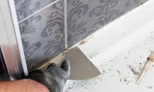Ręka w rękawiczce usuwa stary silikon z narożnika wanny za pomocą szpachelki.