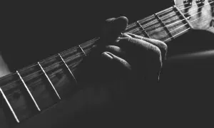 Dłoń dociska struny gitary, przygotowując się do kolejnego akordu. Ciemne tło podkreśla skupienie na grze na gitarze.