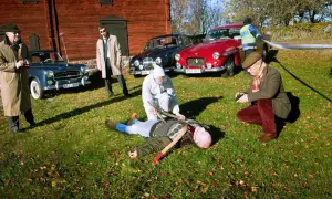 En brottsplats med en död man på marken, omgiven av gamla bilar, inklusive en British Leyland. Polis och utredare arbetar.