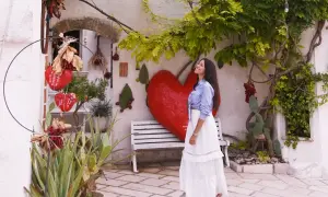 Una donna sorride sotto un albero rigoglioso, con un grande cuore rosso e decorazioni che ricordano il pomodoro ruffano.