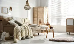 Przytulny salon w stylu hygge wnętrza. Beżowa kanapa z grubym, plecionym kocem, wiklinowa lampa, parawan i świece tworzą ciepłą atmosferę.