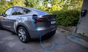 Grå elbil laddas med blå kabel från laddbox på väggen. En grönskande häck syns i bakgrunden.