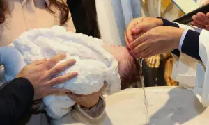 Chrzest święty: ksiądz polewa głowę dziecka wodą. Rodzice trzymają maluszka w białym kocyku.