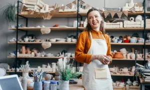 Uśmiechnięta artystka w fartuchu w pracowni ceramicznej, otoczona półkami z wyrobami. Zastanawia się, jak wycenić rękodzieło, by docenić swój trud.