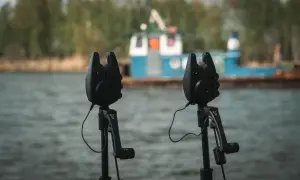 Dwa sygnalizatory brań na grunt gotowe do akcji nad wodą, z łodzią w tle.