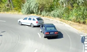 Niebieski samochód wyprzedza białe kombi na zakręcie, ryzykując wymuszenie pierwszeństwa i mandat.