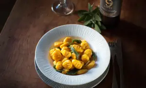Gnocchis de potimarron aux feuilles de sauge, servis dans une assiette blanche sur une table en bois. Une bouteille de vin et un verre sont visibles en arrière-plan. Parfait pour une recette gnocchi potimarron.