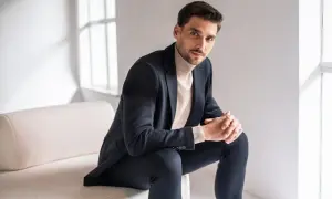 Mężczyzna w granatowym garniturze i beżowym golfie, prezentujący styl smart casual. Siedzi na kanapie, opierając dłonie na kolanach.