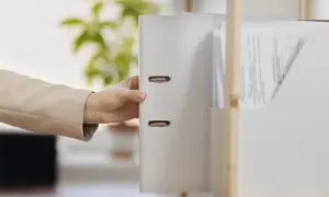 Dłoń w beżowej marynarce sięga po biały segregator z dokumentami. W tle zielone liście, nip-8.