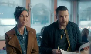 En kvinna med scarf och en man med skägg sitter vid ett bord. Scenen är från en dansk serie på SVT.