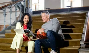 En kvinna och en man sitter på en trappa. Hon läser en tidning, han håller en basketboll. Kanske funderar de på att byta jobb eller inte.