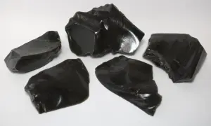 Fünf scharfkantige Stücke glänzenden, schwarzen Obsidiansteins liegen auf weißem Untergrund.