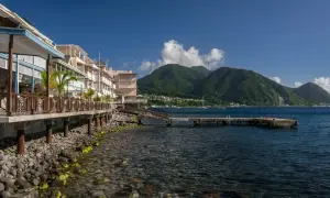 Bâtiments colorés en bord de mer sur l'île Dominique, avec des montagnes verdoyantes en arrière-plan et un quai s'avançant dans l'eau bleue.