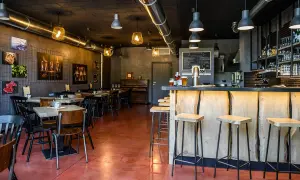 Interno di un locale con tavoli, sedie, un bancone in legno grezzo e sgabelli. Sul bancone due birre, pronte per essere gustate dal baldo birraio.