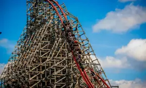 Pasażerowie Hyperion roller coaster zjeżdżają po drewnianej konstrukcji, która wznosi się ku błękitnemu niebu z chmurami.