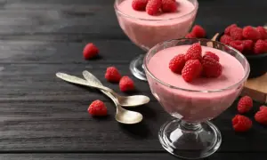 Två glas med hallonmousse, toppade med färska hallon. Två skedar ligger bredvid på ett mörkt träbord.