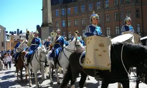 Livgardets dragonmusikkår marscherar i parad. Musiker i blå uniformer spelar på instrument och rider på vita hästar.