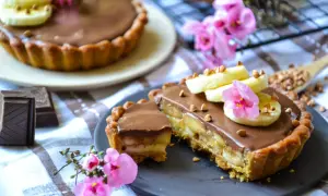 Tarte banane chocolat, garnie de rondelles de banane, de fleurs roses et de noisettes concassées. Un délice gourmand !